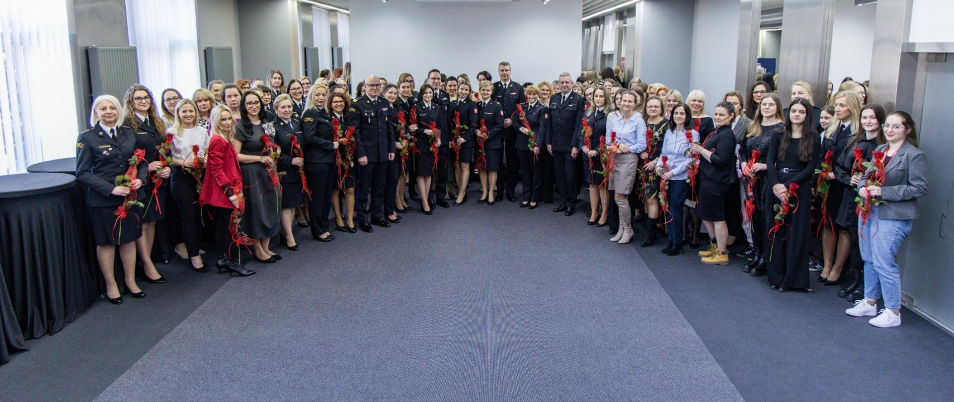 Wspólne zdjęcie uczestniczek wydarzenia oraz kierownictwa Państwowej Straży Pożarnej w sali konferencyjnej Komendy Głównej PSP. Wiele osób trzyma czerwone róże otrzymane podczas uroczystości.