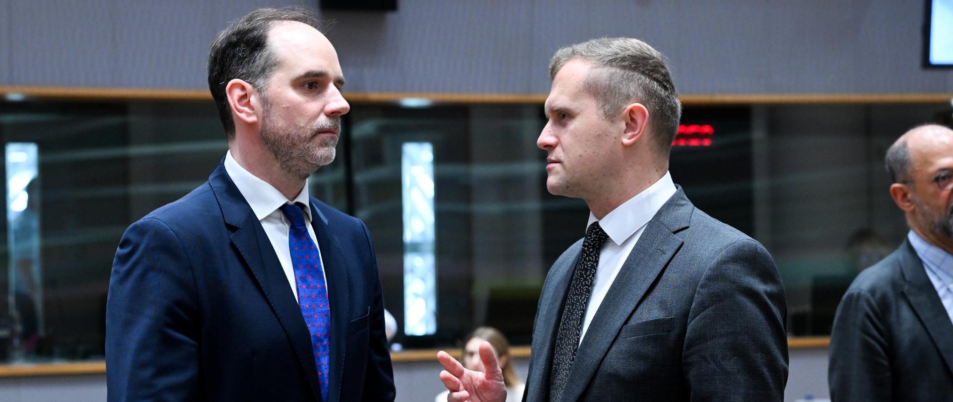 Secretary of State Ignacy Niemczycki and another man are talking in a conference room during an official meeting. Minister Niemczycki is wearing a navy suit with a blue tie, while the other wears a gray suit with a black tie and gestures while speaking. In the background, conference tables, meeting participants, and the modern interior of the room are visible.
