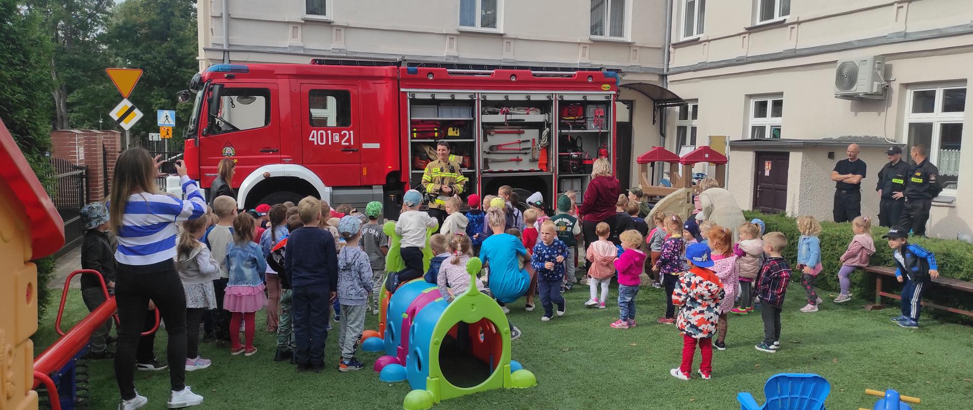 Na zdjęciu widać gromadkę dzieci oraz strażaków, którzy opowiada o pracy. Na dalszym planie samochód strażacki koloru czerwonego. W tle budynek przedszkola.