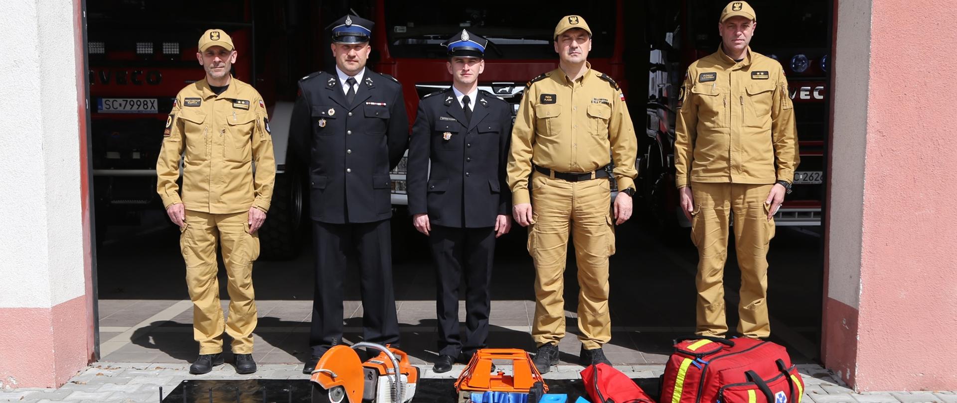 Komendant Centralnej Szkoły PSP w towarzystwie dwóch druhów OSP oraz dwóch oficerów CS PSP, przed nimi mata na której leży przekazywany sprzęt tj: piła do stali i betonu STIHL TS 40, 6 radiotelefonów Motorola GP 340, deska ratownicza, zestaw szyn Kramera oraz torba PSP R1