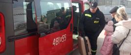 Zdjęcie przedstawia ludzi wsiadających do strażackiego busa. Na pierwszym planie strażak pomagający wsiadać podróżnym.