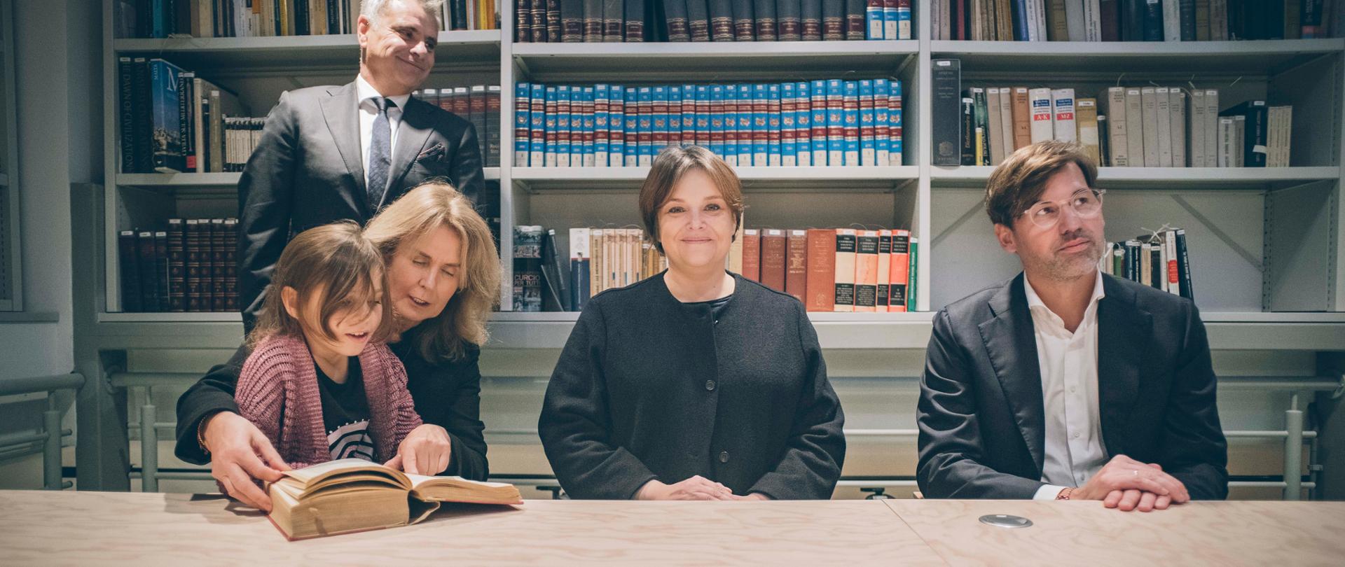 Wiceminister Zioło-Pużuk siedzi za stołem pomiędzy kobietą trzymającą na kolanach małą dziewczynkę a mężczyzną w czarnej marynarce, za nimi półki pełne książek, z lewej stoi mężczyzna w garniturze.