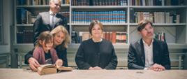 Wiceminister Zioło-Pużuk siedzi za stołem pomiędzy kobietą trzymającą na kolanach małą dziewczynkę a mężczyzną w czarnej marynarce, za nimi półki pełne książek, z lewej stoi mężczyzna w garniturze.