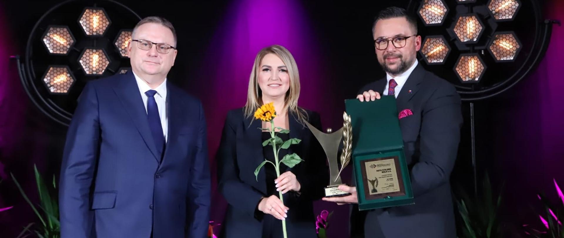 Na zdjęciu wiceminister Marcin Kulasek, kobieta i mężczyzna. Wiceminister trzyma w dłoniach statuetkę, a kobieta słonecznik. W tle kwiaty i dwa reflektory. 