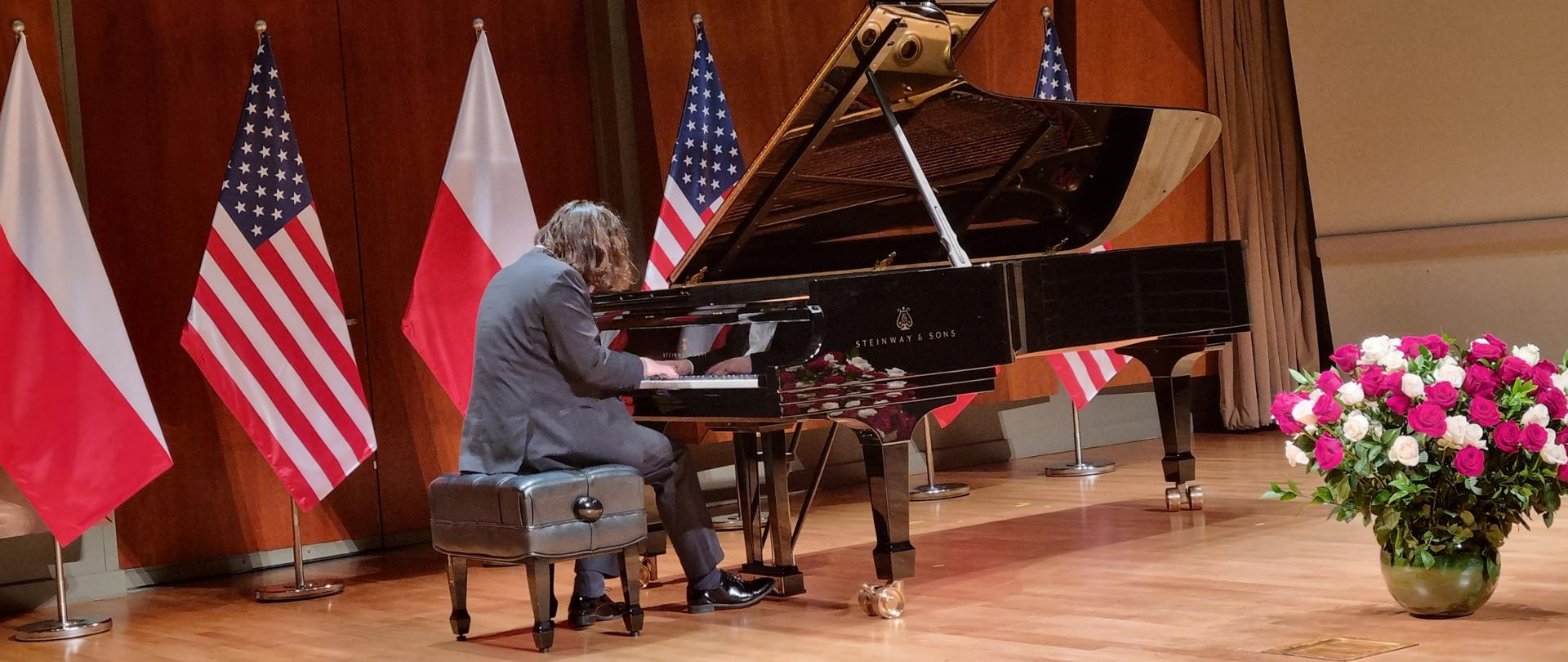 Piano recital by Jakub Kuszlik on the occasion of Poland's National Independence Day. The pianist performs works by Polish composers on a black king on stage at the Colburn School in Los Angeles. In the background are the flags of Poland and the United States. A red and white bouquet of roses stands in front of the piano. 
