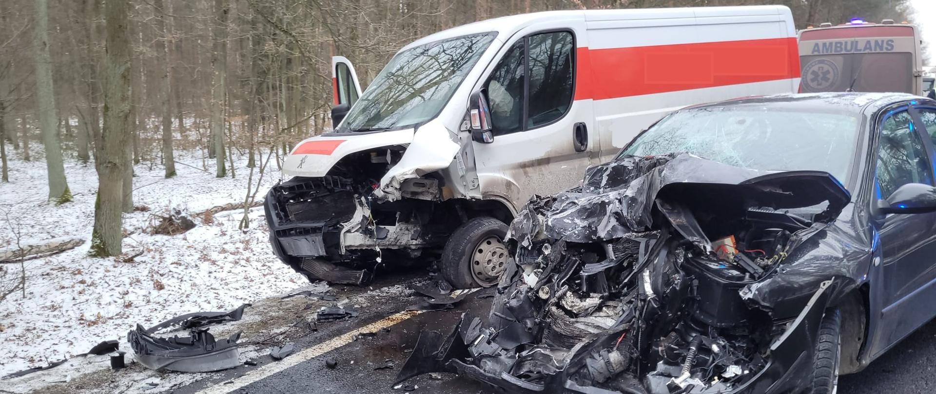 Na wąskiej drodze przebiegającej przez las stoją dwa mocno rozbite pojazdy po zderzeniu czołowym. Czarny samochód osobowy ma całkowicie zgnieciony przód, odsłonięty silnik i powykrzywianą maskę. Biały bus z czerwonym pasem również jest poważnie uszkodzony z przodu. Na mokrym asfalcie widać rozsypane fragmenty karoserii. W tle, między drzewami i przy drodze, stoi karetka. Wokół zalega cienka warstwa śniegu, a drzewa są bez liści, co sugeruje zimową porę.