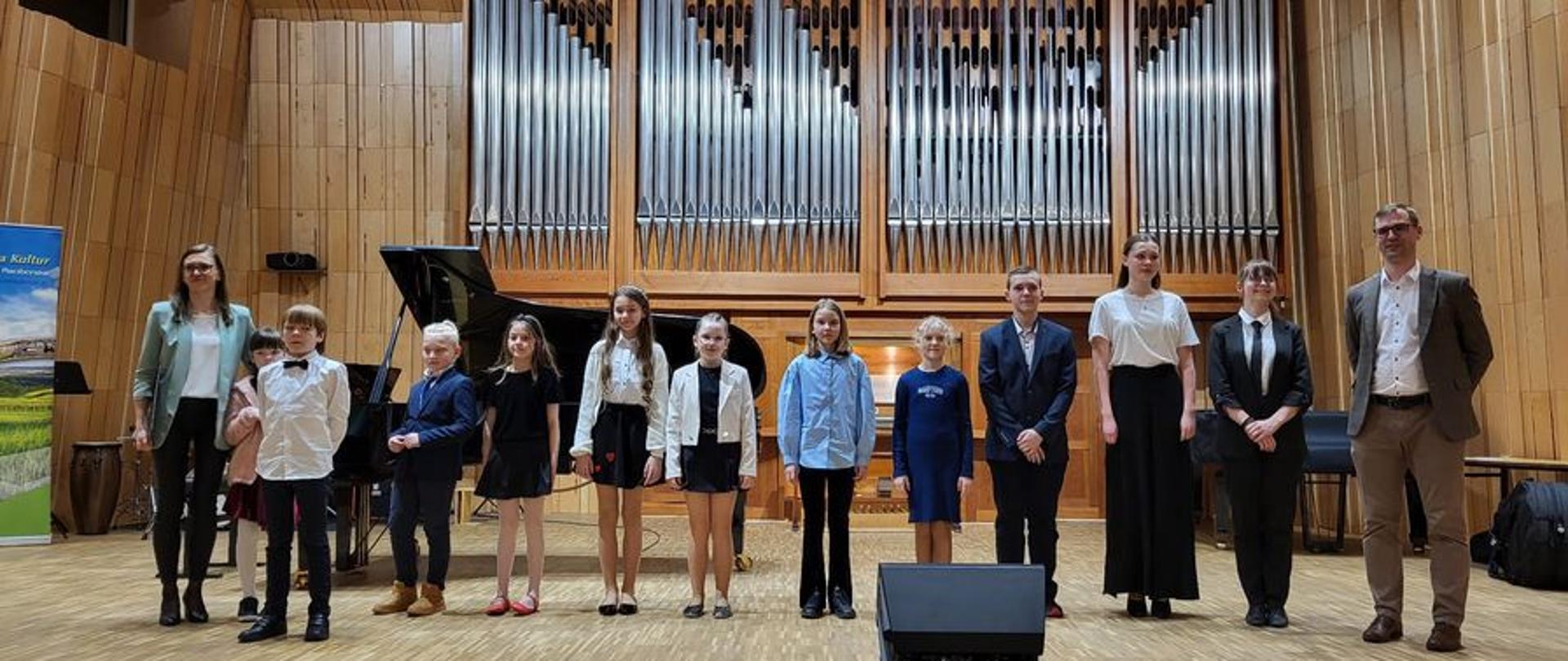 Grupa uczniów i ich nauczyciele stoją na scenie sali koncertowej, w tle widoczny prospekt organów.