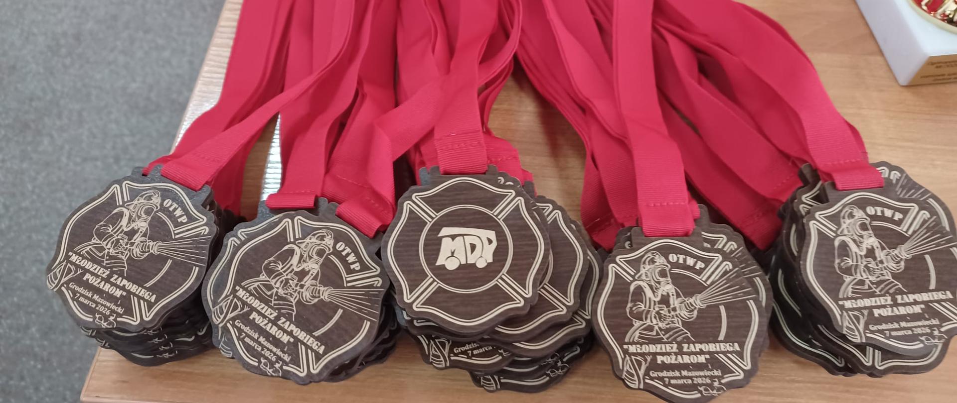 Zbliżenie na drewniane medale w kształcie krzyża maltańskiego z wygrawerowaną postacią strażaka i napisem „Młodzież Zapobiega Pożarom”. Medale mają szerokie, czerwone wstążki i leżą ułożone w stosach na jasnym, drewnianym blacie.