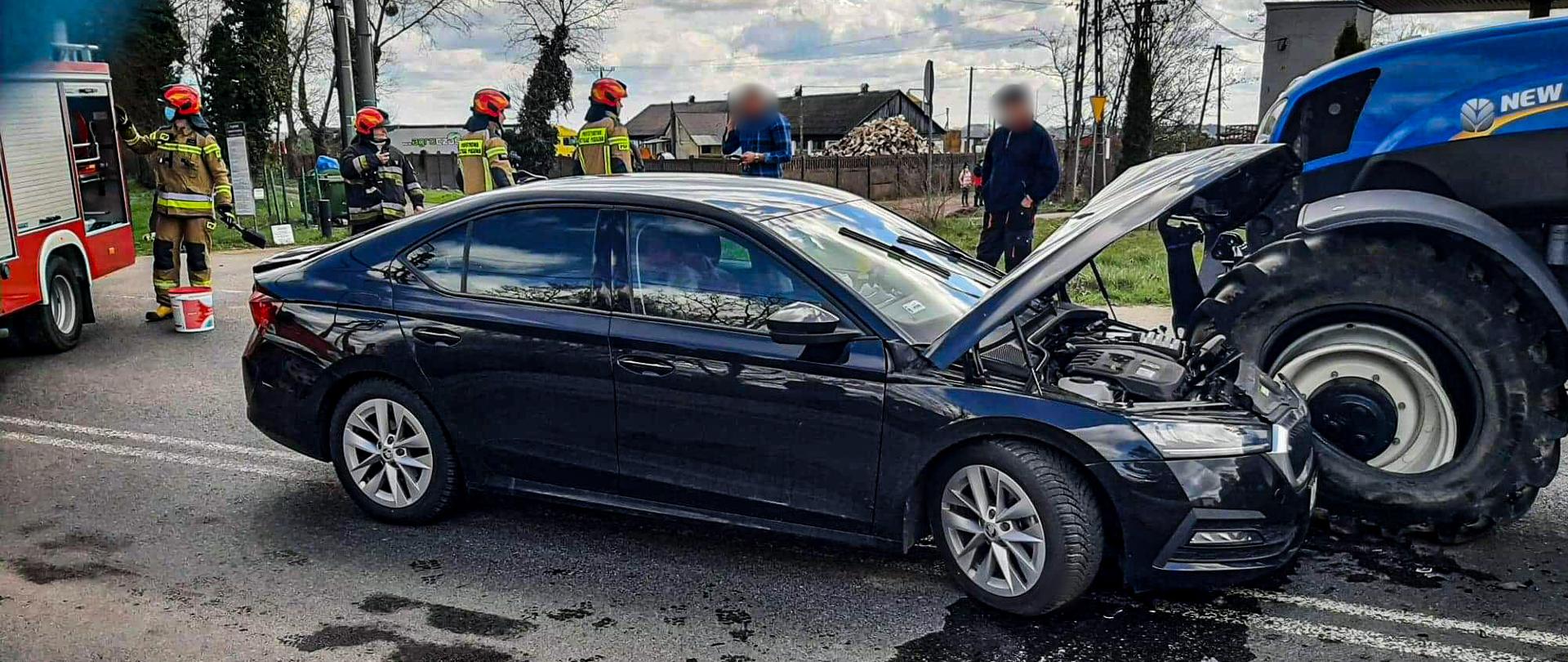 Na zdjęciu w środkowej części znajduje się samochód osobowy z podniesioną maską oraz przód ciągnika rolniczego z przebitym kołem przednim, z tyłu samochodu znajdują się strażacy pobierający sprzęt z wozu strażackiego.
