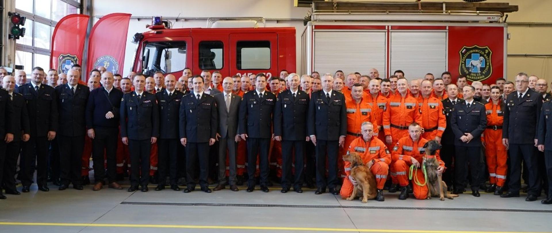 Na zdjęciu wszyscy uczestnicy uroczystości - strażacy PSP w mundurach wyjściowych oraz strażacy w pomarańczowych kombinezonach grupy poszukiwawczo-ratowniczej. Na przodzie zdjęcia wraz z opiekunami widoczne są dwa psy ratownicze. W tle widoczny jest duży czerwony samochód strażacki.