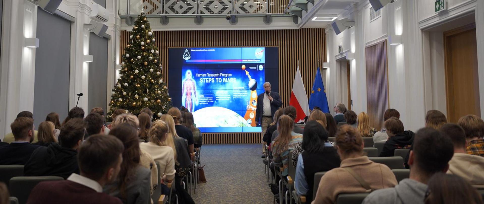 Widok z tyłu na salę, na krzesłach siedzą ludzie, przed nimi na ścianie wielki ekran, obok niego choinka.