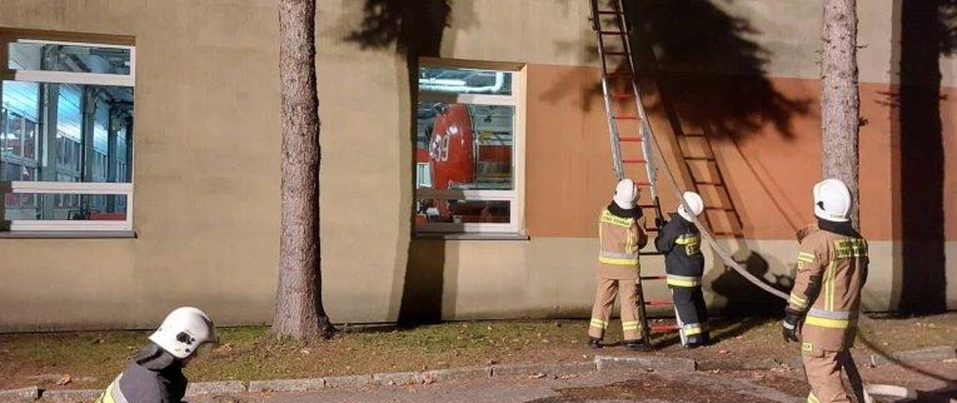 Zdjęcie przedstawia strażaków zabezpieczających drabinę przy ścianie budynku. 