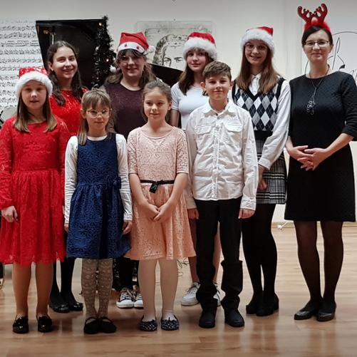 Uczniowie klasy fortepianu stoją wraz z nauczycielem na tle fortepianu. Uczniowie mają na głowach czapki Mikołaja.