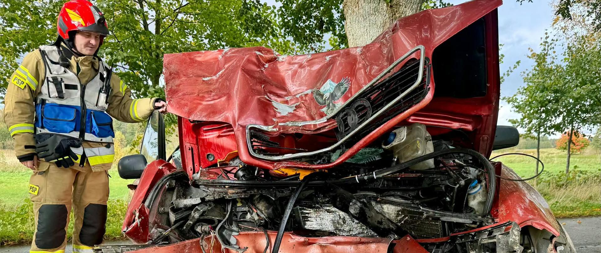 Na zdjęciu widoczny jest strażak Państwowej Straży Pożarnej w ubraniu specjalnym i hełmie, stojący obok poważnie uszkodzonego czerwonego pojazdu. Przód samochodu jest całkowicie zmiażdżony, a elementy karoserii, silnika i wyposażenia są zdeformowane oraz porozrzucane na ziemi. Pojazd znajduje się na poboczu drogi, w otoczeniu drzew i terenów zielonych.