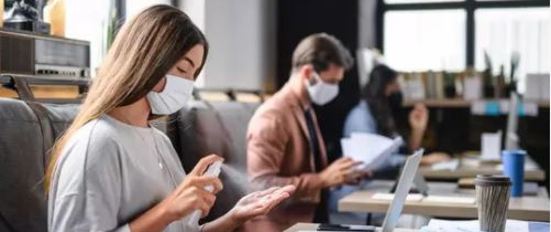Na zdjęciu znajdują się trzy osoby pracujące w maseczkach. Kobieta na pierwszym planie dezynfekuje dłonie. Mężczyzna znajdujący się na drugim planie przegląda dokumenty.