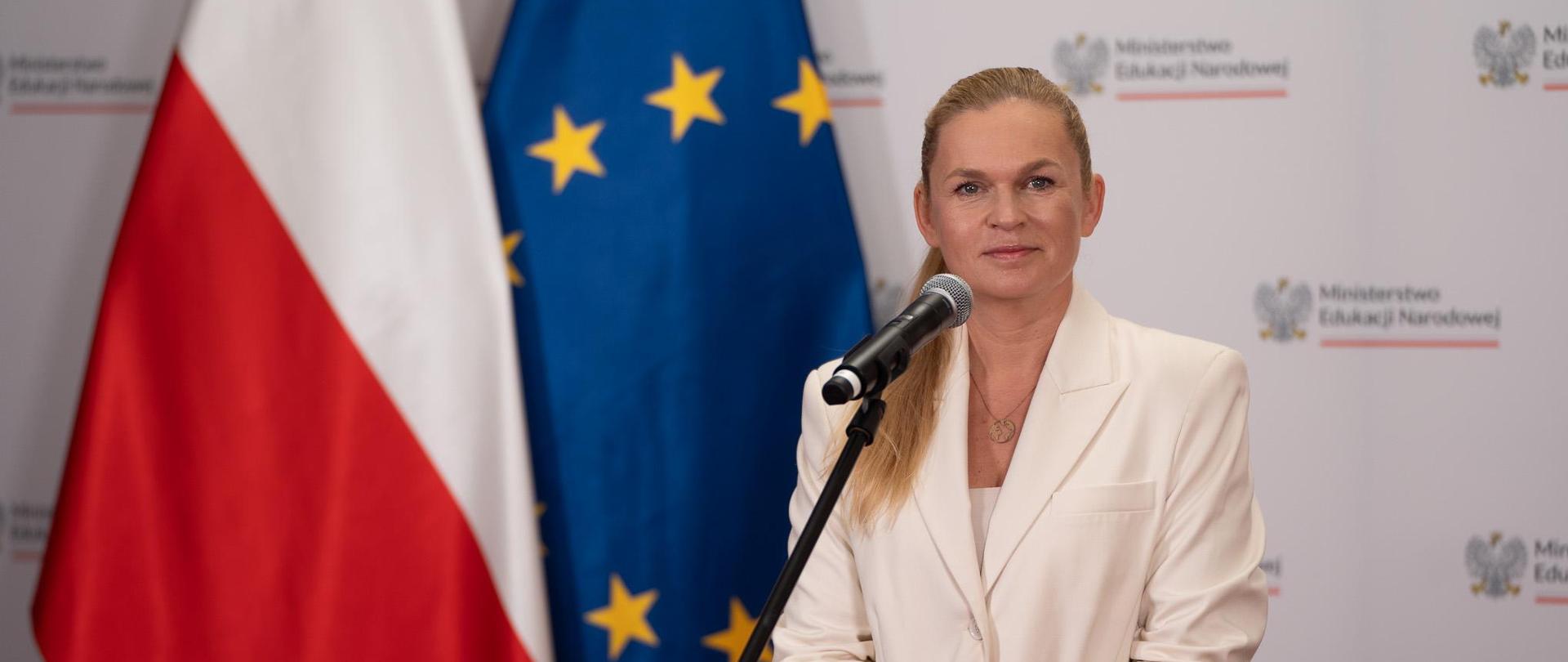 Minister Barbara Nowacka stoi przy mikrofonie i mówi, w tle flagi Polski UE
