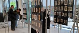 Na pierwszym planie widać ramy wystawiennicze w których wyeksponowane są projekty ekslibrisów, w tle widać zwiedzających, oglądających projekty oraz duże przeszklone ściany okien
