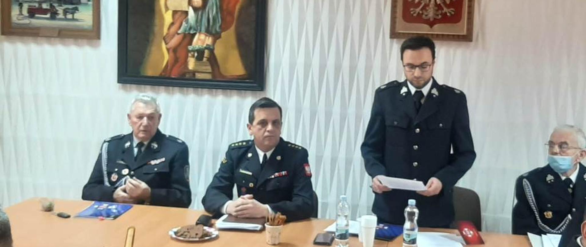 Sala Narad OSP Włoszczowa, stół prezydialny przy którym zasiadają od lewej dh Mirosław Pawlak, st. kpt. Andrzej Pacanowski, dh Bartłomiej Robak (odczytuje sprawozdanie z działalności), dh Wiktor Krotla, w tle ściana z obrazem św. Floriana oraz godło Polski