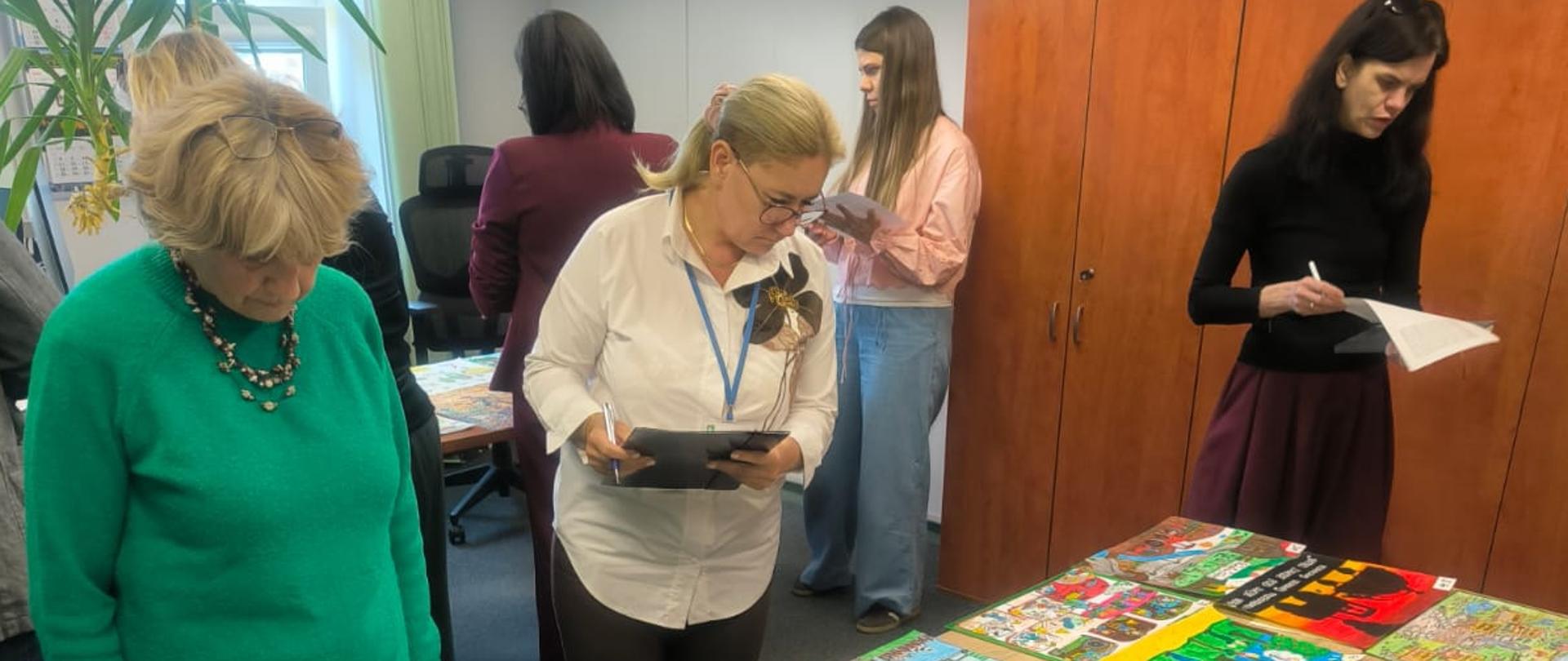 Na środku pokoju, na stole, wyłożonych jest piętnaście prac plastycznych. Wokół stołu kilka osób trzymających w rękach arkusze oceny pochyla się nad pracami dokonując ich oceny 
