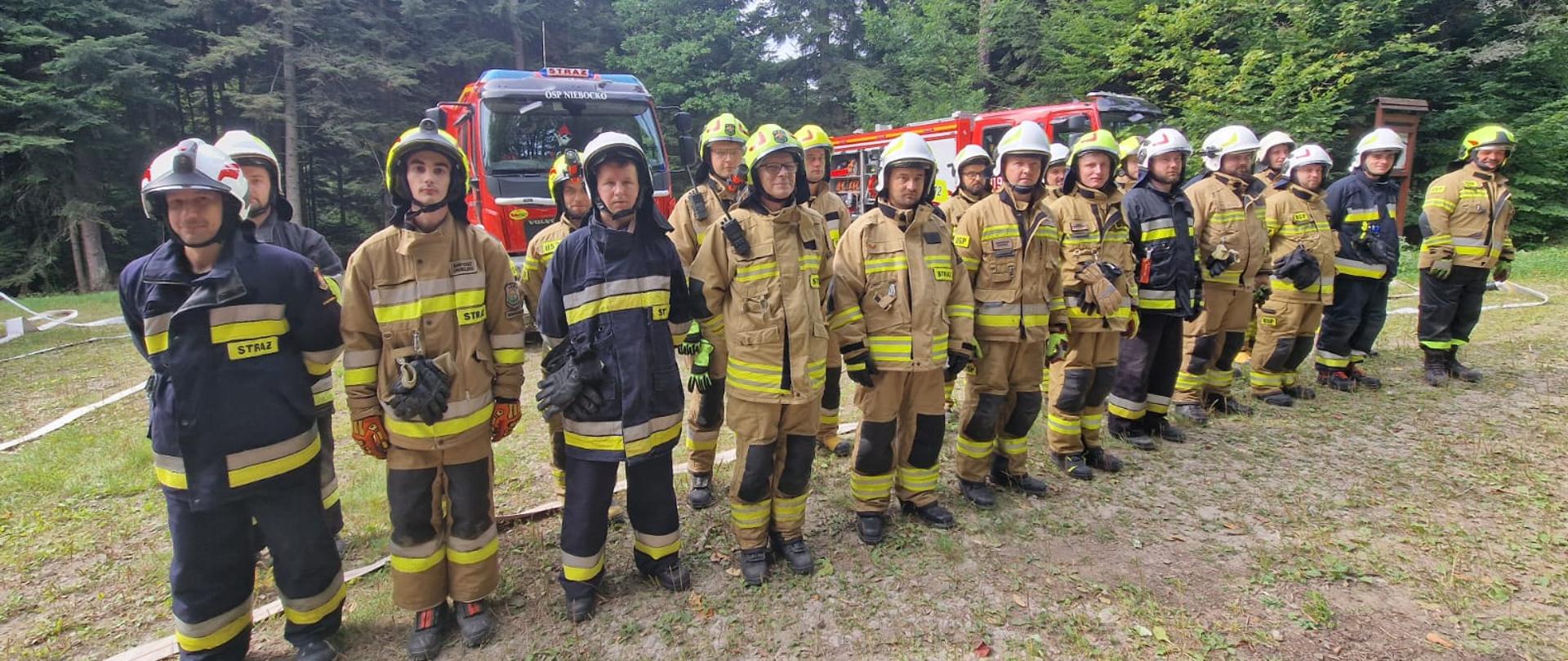 Kolorowa fotografia wykonana w pochmurny dzień w okolicach lasu. Przedstawia strażaków ubranych w ubrania specjalne z hełmami po zakończonym egzaminie praktycznym szkolenia kierowców konserwatorów OSP. W tle samochody pożarnicze biorące udział w egzaminie.