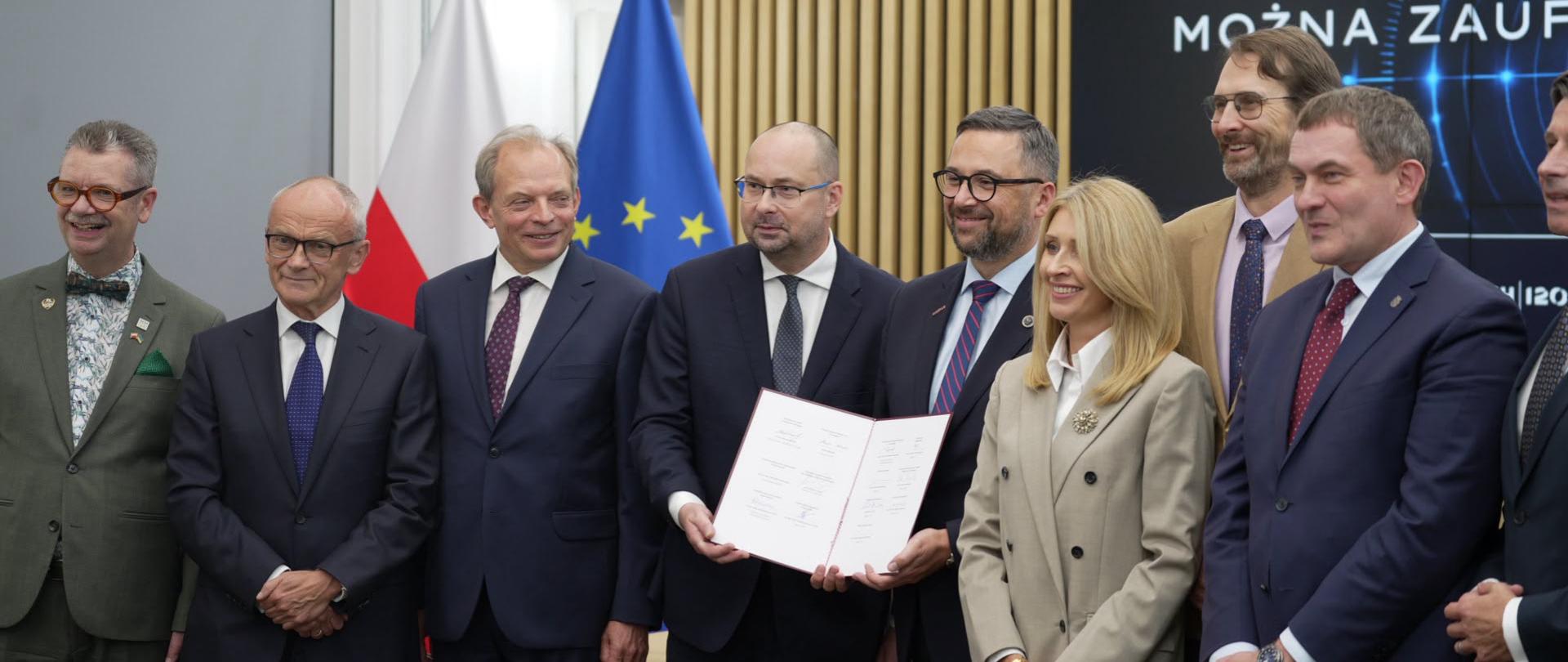 Grupa uczestników wydarzenia stoi do pamiątkowego zdjęcia po podpisaniu porozumienia. W centralnej części kadru dwie osoby prezentują otwarty dokument zawierający podpisy. Pozostali uczestnicy stoją obok w półkolu. W tle widoczne są flagi Polski i Unii Europejskiej, a także duży ekran z logotypem PAP Naukowego Think Tanku oraz hasłem „Wiedza, której można zaufać”. Zdjęcie wykonano w nowoczesnej sali konferencyjnej wykończonej pionowymi drewnianymi panelami.