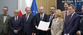 Grupa uczestników wydarzenia stoi do pamiątkowego zdjęcia po podpisaniu porozumienia. W centralnej części kadru dwie osoby prezentują otwarty dokument zawierający podpisy. Pozostali uczestnicy stoją obok w półkolu. W tle widoczne są flagi Polski i Unii Europejskiej, a także duży ekran z logotypem PAP Naukowego Think Tanku oraz hasłem „Wiedza, której można zaufać”. Zdjęcie wykonano w nowoczesnej sali konferencyjnej wykończonej pionowymi drewnianymi panelami.