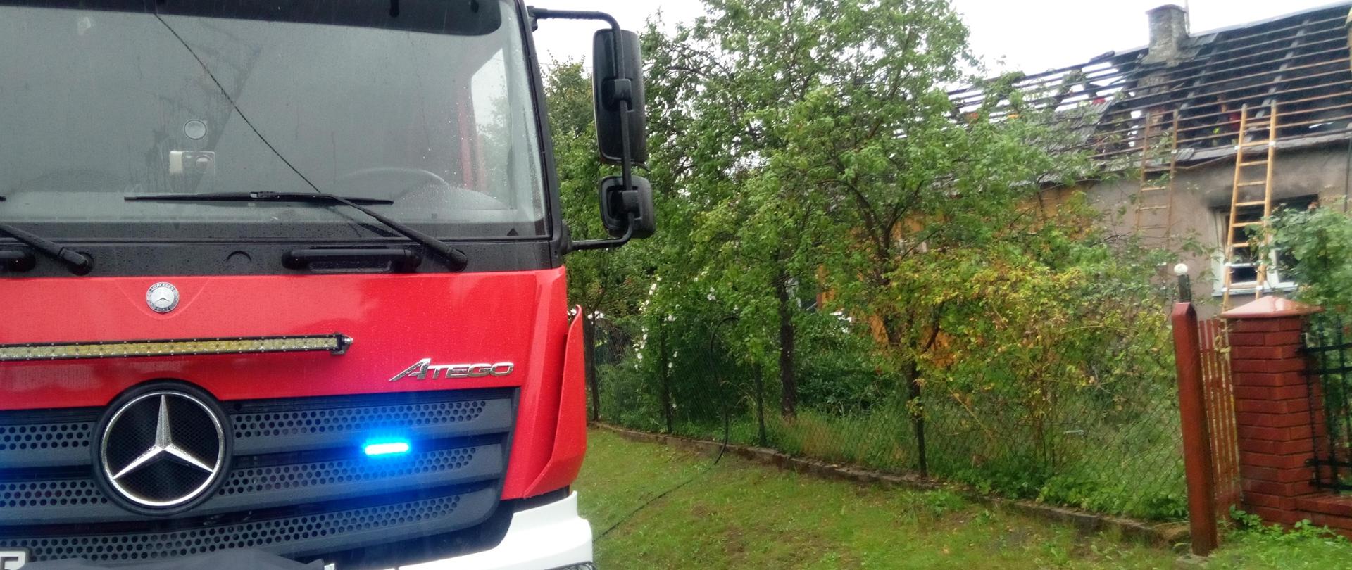 Zdjęcie przedstawia po prawej stronie budynek jednorodzinny, który został zniszczony przez pożar, po lewej samochód pożarniczy. Widać ogrodzenie posesji i odsłoniętą, zniszczoną przez ogień więźbę dachową. 