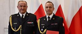 Dwóch strażaków w mundurach wyjściowych ze sznurem stoi obok siebie jeden z mężczyzn trzyma czerwoną teczkę za nimi ustawione są trzy flagi Polski oraz znajdują się drzwi. 