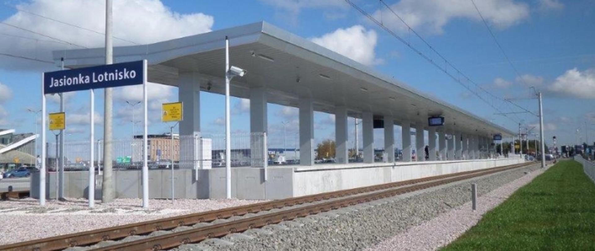 Railway station platform with the sign "Jasionka Lotnisko," featuring a roof supported by columns, tracks alongside, and background buildings under a partly cloudy sky.