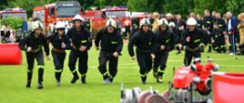 Na zielonej trawie na podeście stoi czerwona motopompa a obok niej czerwone i białe węże pożarnicze, w niedalekiej odległości za motopompą widać 7 strażaków, którzy rozpoczynają bieg, są ubrani w czarne mundury i mają białe hełmy na głowie. W tle widać grupę obserwatorów, większość z nich ubrana jest w mundury strażackie, widać również czerwony samochody strażackie.