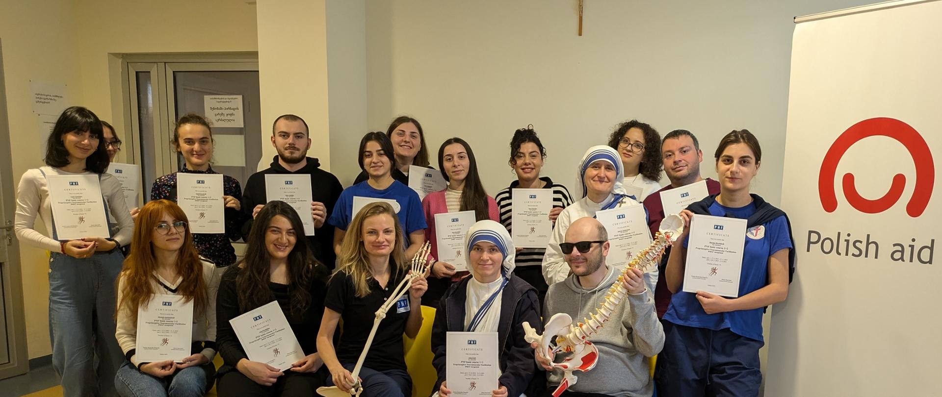 Grupa kobiet i mężczyzn pozuje z certyfikatami, z przodu siedzą dwie osoby, z których jednak ma model szkieletu dłoni a druga szkieletu kręgosłupa i miednicy