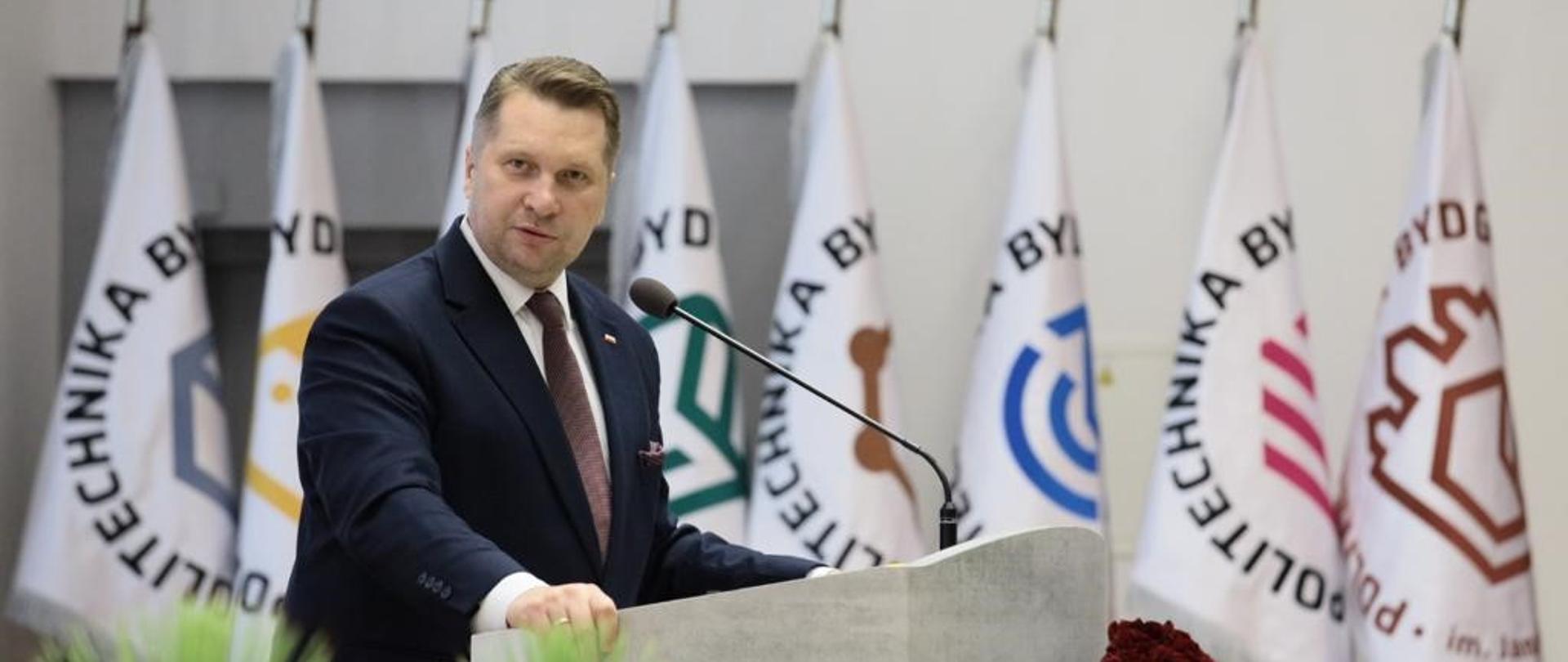 Minister Przemysław Czarnek stoi przy mównicy. Ma lekko odchyloną głowę. Jest ubrany w garnitur i brązowy krawat. Na pierwszym planie kompozycja kwiatowa. W tle białe flagi z logotypami Politechniki Bydgoskiej