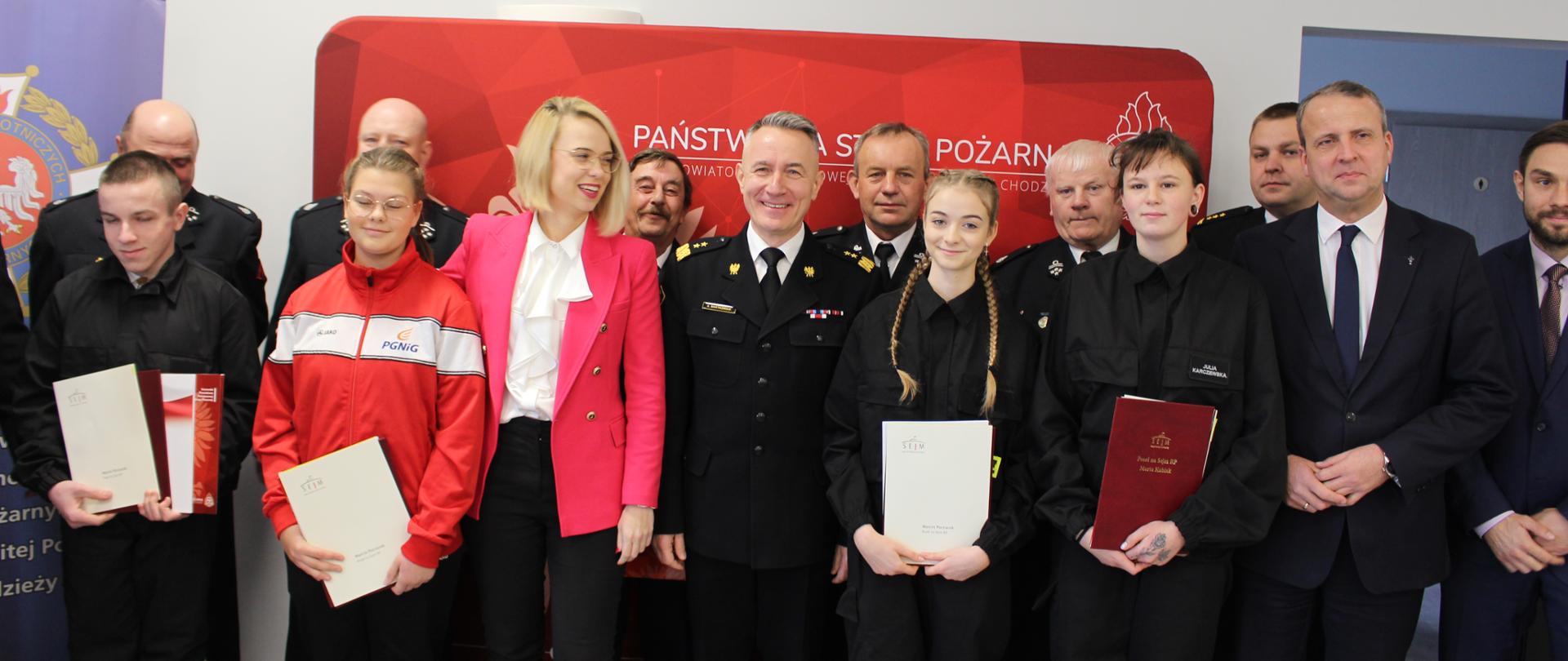 zdjęcie grupowe strażaków oraz młodzieży w strojach strażackich , zdjecie wykonane na czerwonym tle 