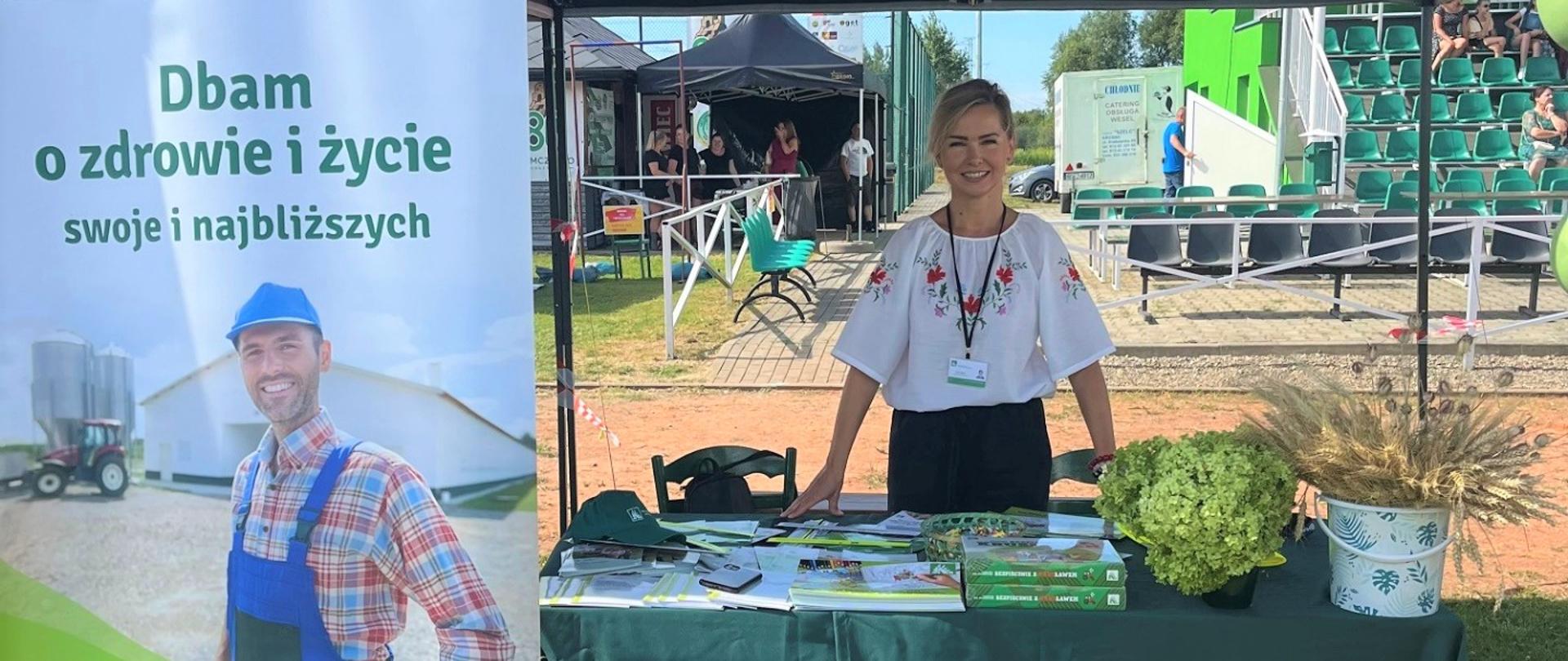 Przy stoisku informacyjno-promocyjnym Kasy znajduje się jedna kobieta – pracownica KRUS. Przed nią znajduje się stół, na którym umieszczone są materiały prewencyjne Kasy oraz kwiaty. Po lewej stronie znajduje się roll-up z hasłem: „Dbam i zdrowie i życie swoje i najbliższych”.