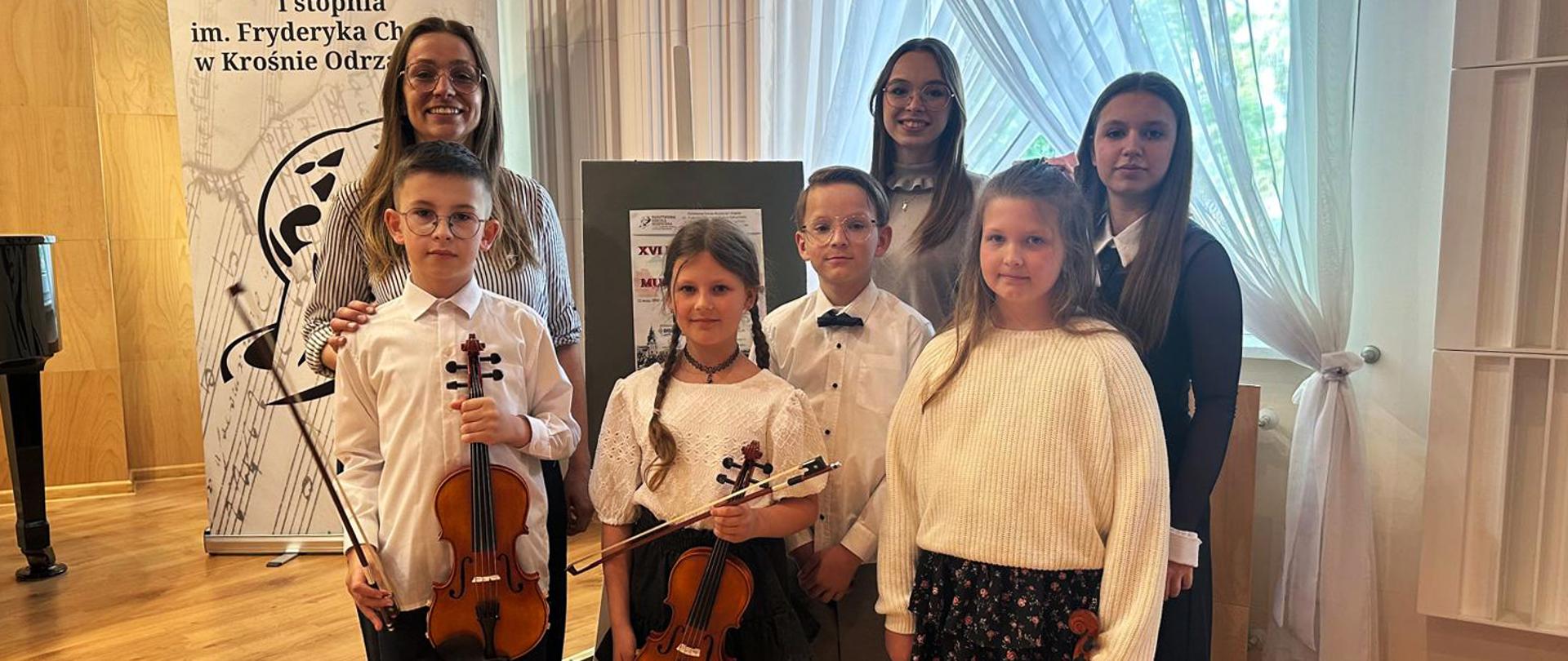 Na zdjęciu dwie nauczycielki i czworo uczniów stoją w w auli szkoły. Dwoje dzieci trzyma w ręku skrzypce.