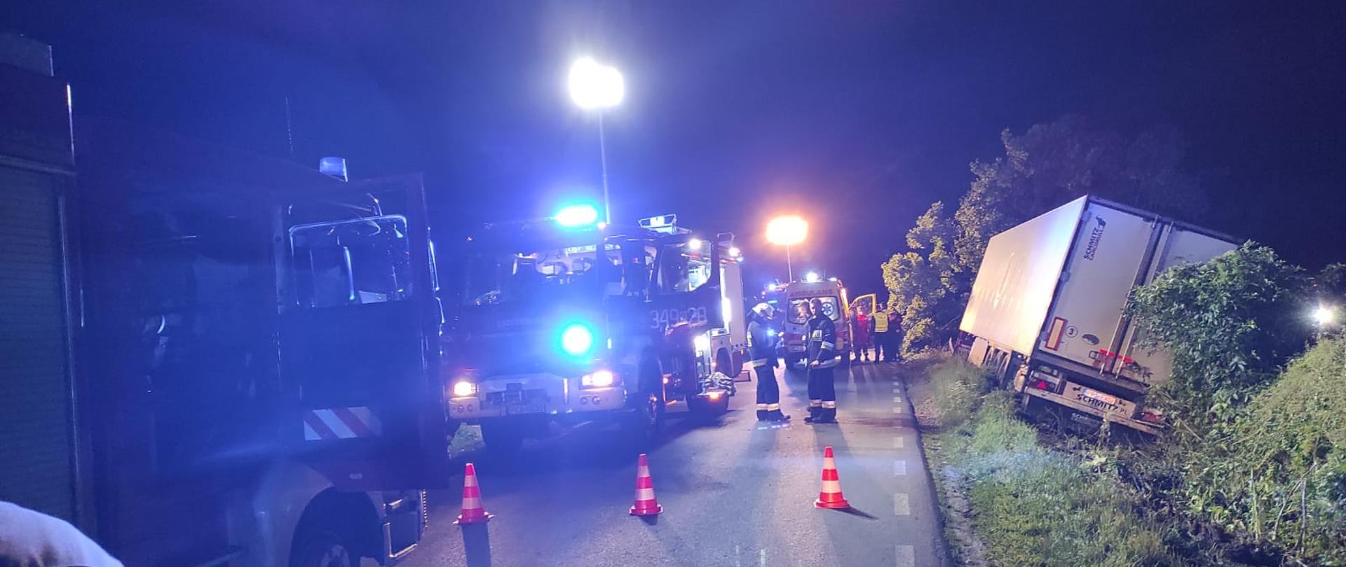 Akcja ma miejsce w nocy. Na zdjęciu z prawej strony kadru widzimy tył samochodu ciężarowego koloru białego, który stoi przechylony na prawą stronę w rowie. Do samochodu znajdującego się w rowie przylegają krzaki oraz zarośla koloru zielonego. Środek oraz lewa strona zdjęcia droga asfaltowa na której stoją służby ratunkowe, dwa samochody strażackie oraz karetka pogotowia. Samochody strażackie koloru czerwonego, widzimy niebieskie światła ostrzegawcze. Obok samochodów strażackich rozstawione na asfalcie trzy pachołki czerwono białe. Dalej widzimy karetkę oraz ratowników medycznych. 