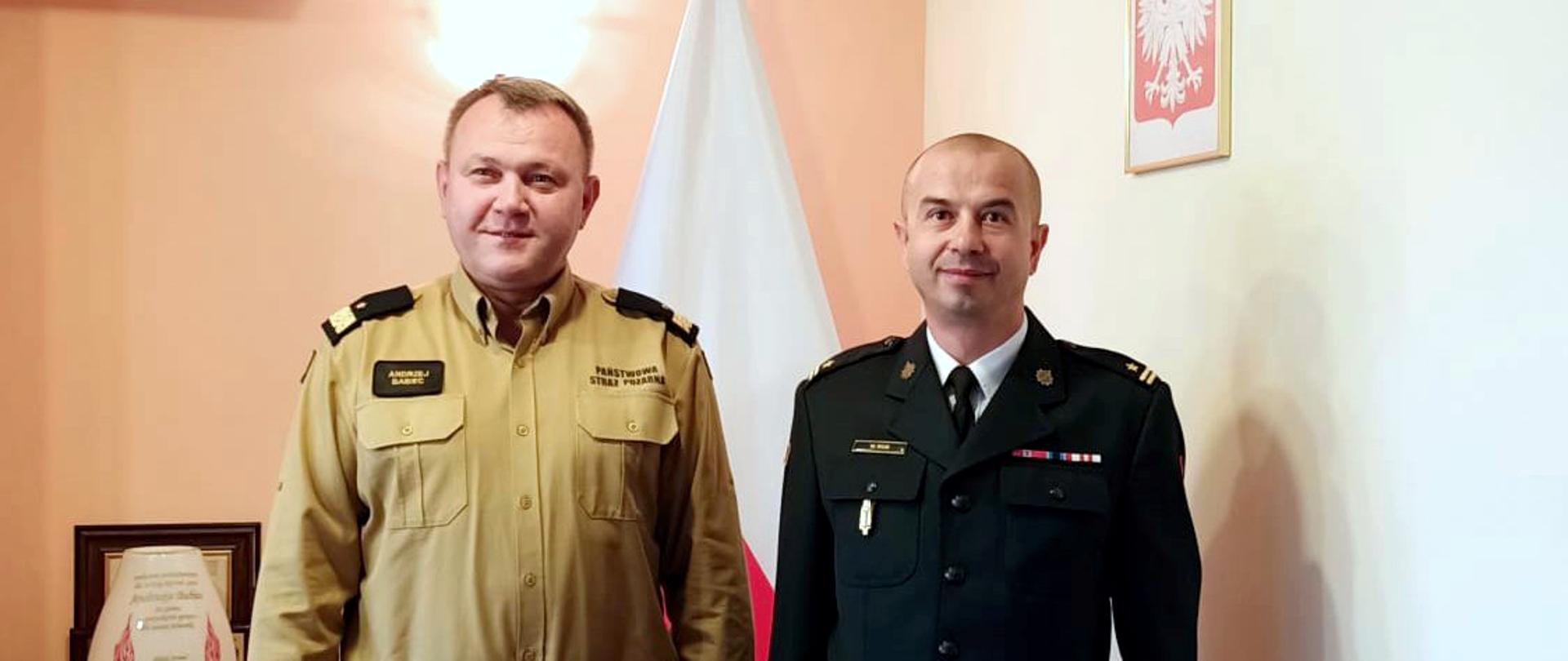 Zdjęcie zrobione w pomieszczeniu. W szeregu, w umundurowaniu wyjściowym stoi dwóch funkcjonariuszy Państwowej Straży Pożarnej. Z lewej strony funkcjonariusz w stopniu generała, z prawej w stopniu młodszego brygadiera