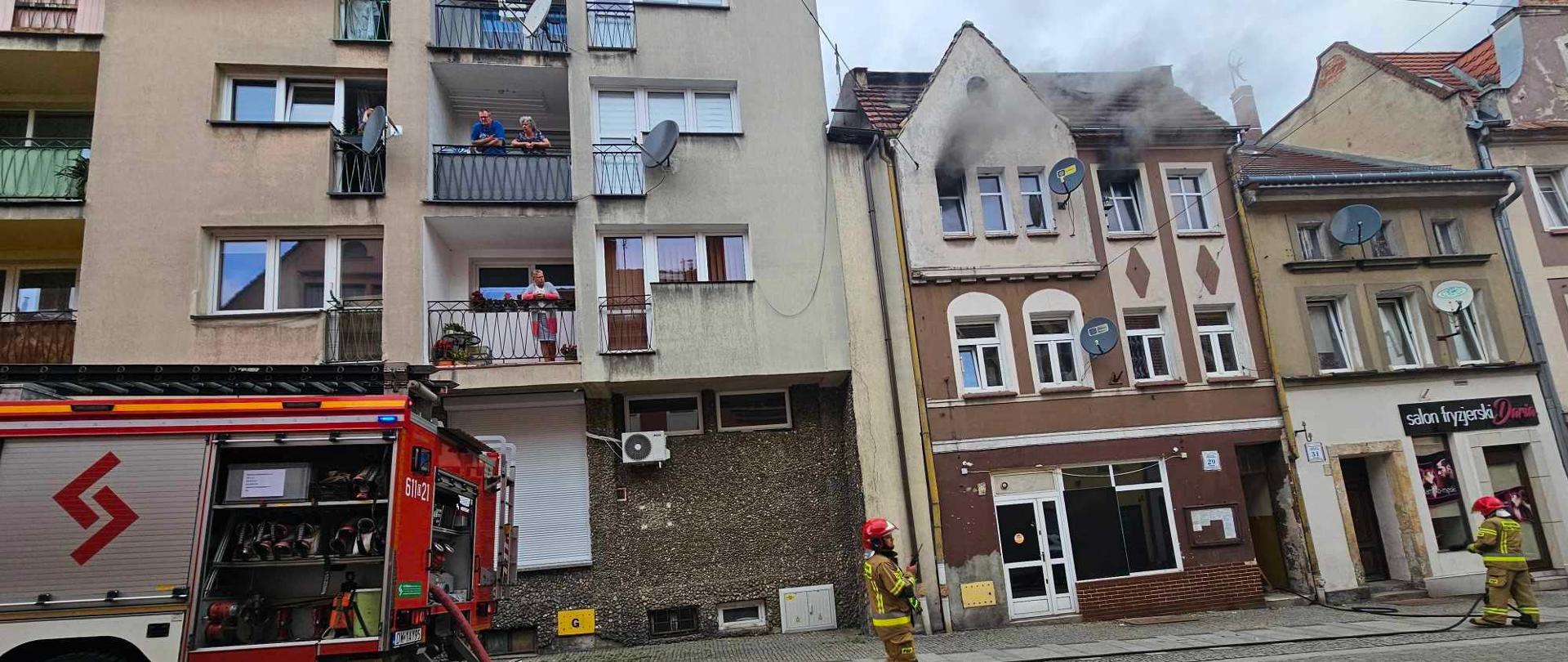 droga w rynku pomiędzy kamienicami, strażacy rozwijają linię gaśniczą z pojazdu, z okien kamienicy wydobywa się dym