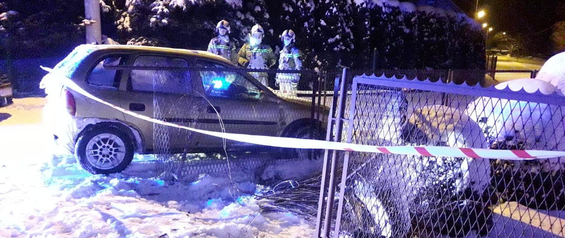 Inne ujęcie samochodu stojącego obok zniszczonego ogrodzenia. W tle stoi trzech strażaków. Wokół leży śnieg.