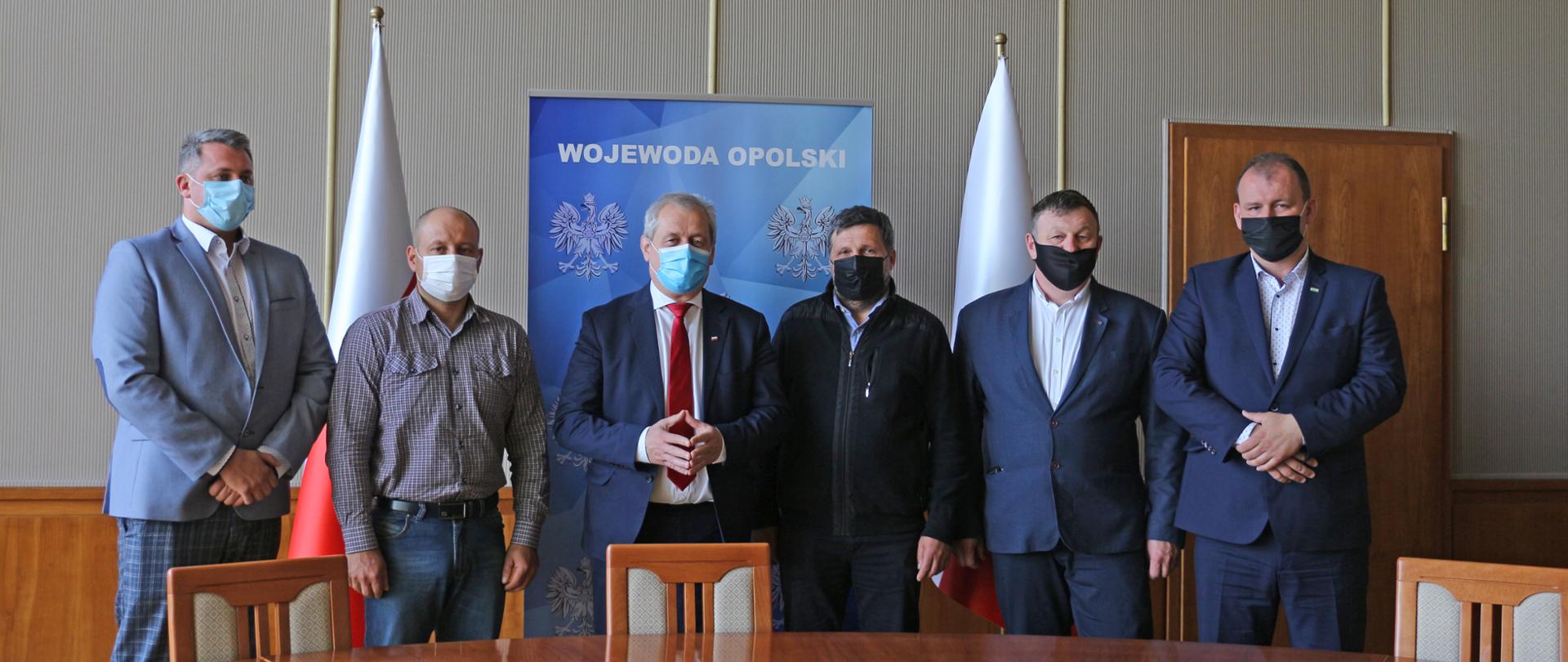 Na zdjęciu 6 mężczyzn, stojących obok siebie i pozujących do wspólnego zdjęcia. Mężczyźni stoją na tle roll upu z napisem Wojewoda Opolski oraz dwóch biało-czerwonych flag, które są po boku. Przed mężczyznami widać fragment blatu okrągłego stołu, w jasnym kolorze. Za stołem 3 drewniane krzesła. W tle ściana oraz - w górnej części godło. W środku mężczyzn stoi wojewoda opolski. 