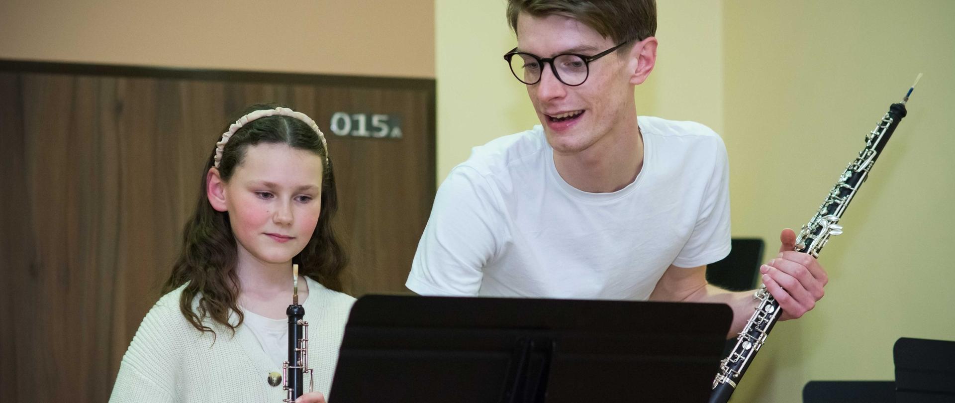 zdjęcie przedstawia prowadzącego warsztaty trzymającego w lewej dłoni obój, obok uczennica trzymająca obój, na pierwszym planie pulpit, prowadzący udziela wskazówek