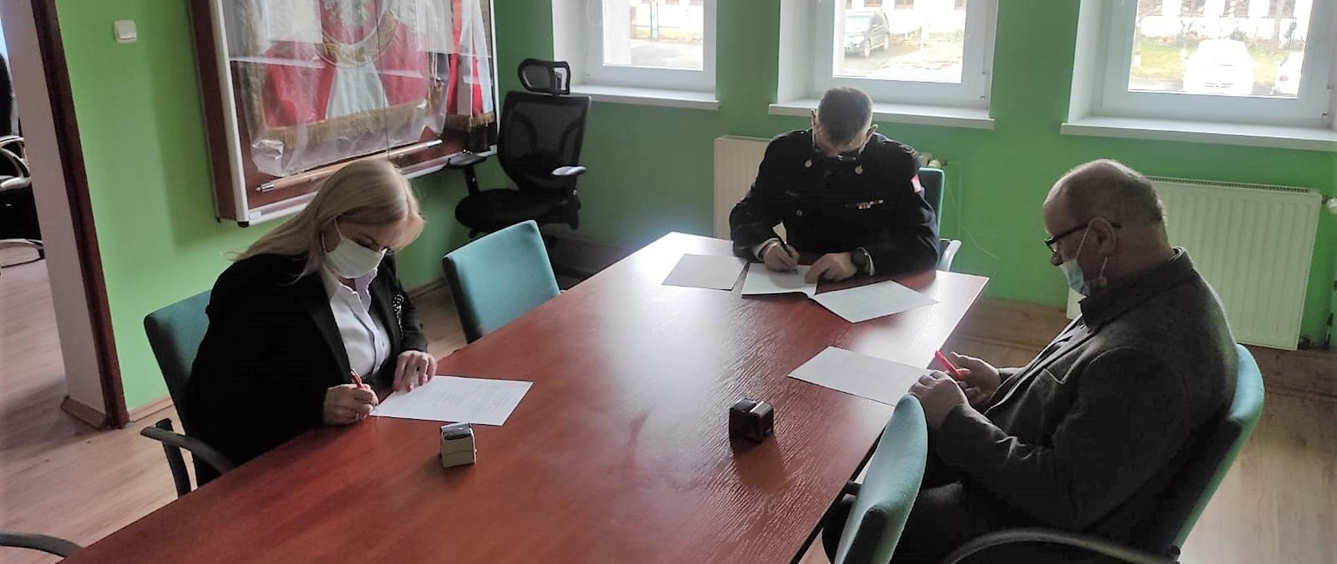 Obraz przedstawia Komendanta Powiatowego brygadiera Waldemara Sarleja, Wójt Gminy Wądroże Wielkie panią Elżbietę Jedlecką oraz Prezesa OSP Budziszów Wielki druha Zdzisława Osaka podpisujących porozumienie o włączeniu jednostki do krajowego systemu ratowniczo-gaśniczego. Osoby w sali narad. Pora dzienna.