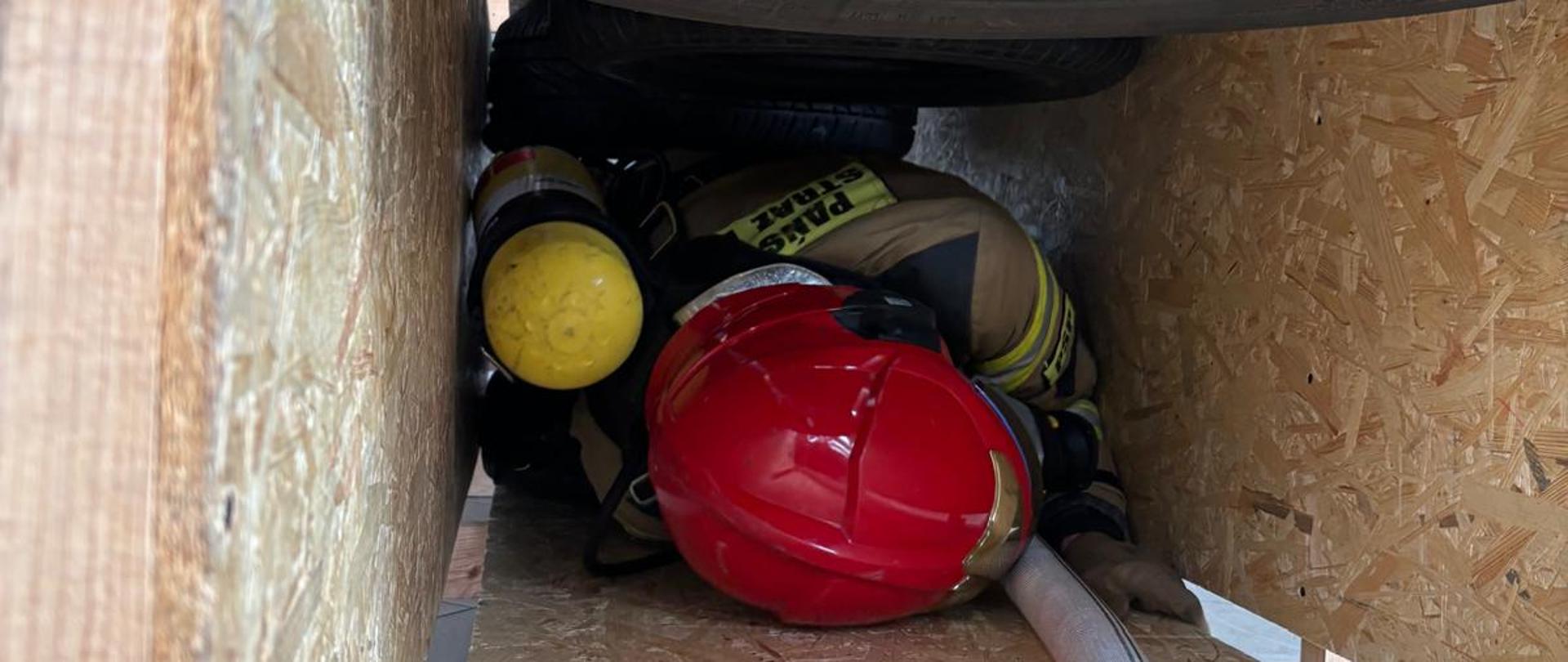 strażak w ciasnym otworze czołga się, nad nim wisi opona, na po dole wąż pożarniczy 