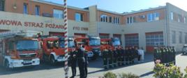 Poczet flagowy stoi przed masztem i przyczepia flagę za nimi stoją strażacy w mundurach koszarowych oraz pracownicy cywilni w rzędach naprzeciwko siebie za nimi wystawione są cztery samochody gaśnicze.