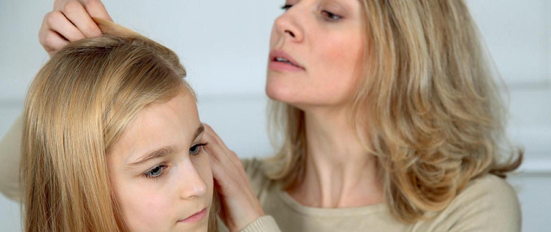 Na zdjęciu widzimy kobietę oraz dziewczynkę w wieku szkolnym. Kobieta sprawdza włosy dziewczynki. Całość przedstawiona jest w łagodnych jasno-beżowych barwach.