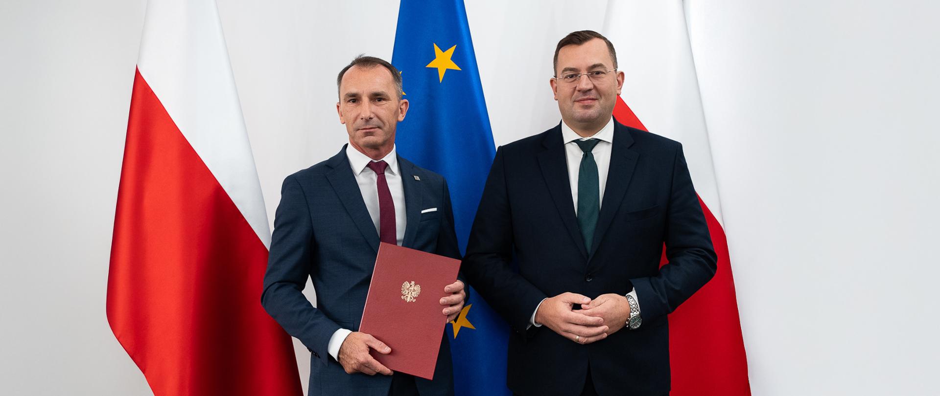 Na tle dwóch polskich flag biało-czerwonych i jednej niebieskiej flagi Unii Europejskiej stoją dwaj mężczyźni. Uśmiechają się do zdjęcia. Mężczyzna stojący z lewej trzyma w rękach bordową ozdobną teczkę z ze złotym godłem Polski.