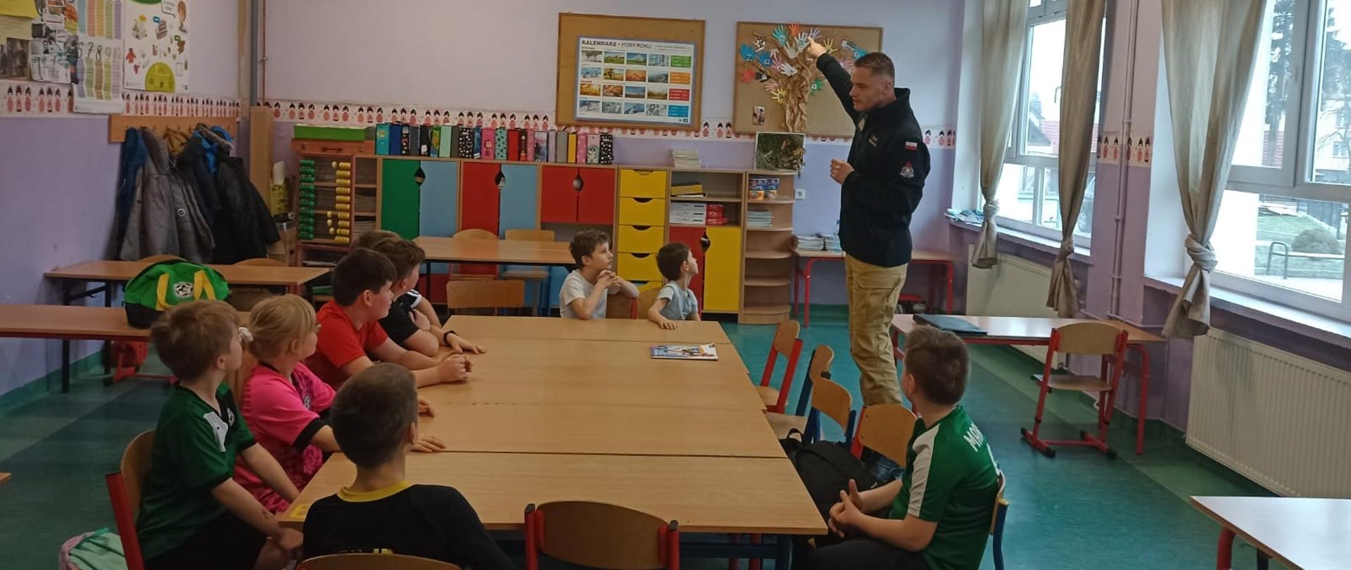 Zdjęcie przedstawia funkcjonariusza Komendy Powiatowej Państwowej Straży Pożarnej w Krośnie Odrzańskim na tle uczestników ferii zimowych, który prowadzi pogadankę na temat zasad bezpieczeństwa podczas zimowego wypoczynku.