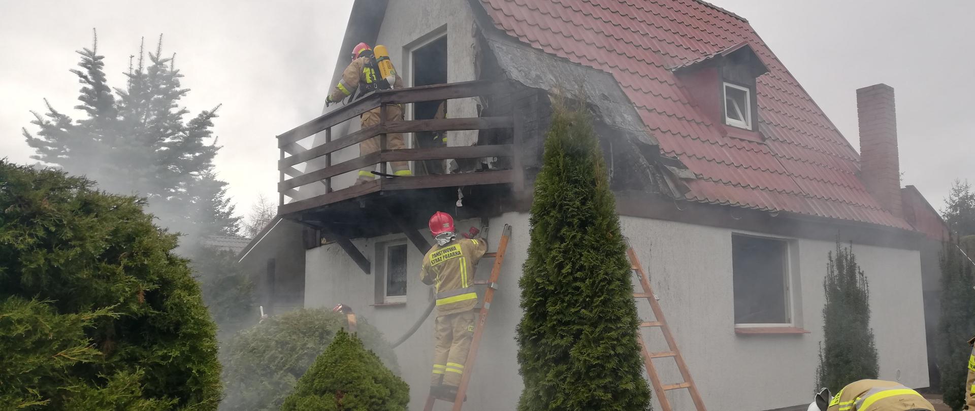 Na zdjęciu widać lekko zadymiony domek letniskowy z poddasze i dachem spadzistym . Dolny narożnik domku o wymiarach około 2mx2m spalony. Na balkonie stoi strażak w aparacie oddechowym natomiast o ścianę budynku oparte są dwie drabiny przystawne. Na jednej z nich stoi strażak z wężem pożarniczym