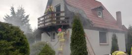 Na zdjęciu widać lekko zadymiony domek letniskowy z poddasze i dachem spadzistym . Dolny narożnik domku o wymiarach około 2mx2m spalony. Na balkonie stoi strażak w aparacie oddechowym natomiast o ścianę budynku oparte są dwie drabiny przystawne. Na jednej z nich stoi strażak z wężem pożarniczym