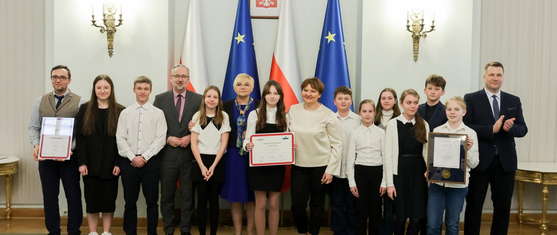 Pod białą ścianą na której wiszą ozdobne kinkiety stoi grupa ubranych na galowo młodych ludzi, troje trzyma oprawione w ramki dokumenty, za nimi flagi Polski i UE.